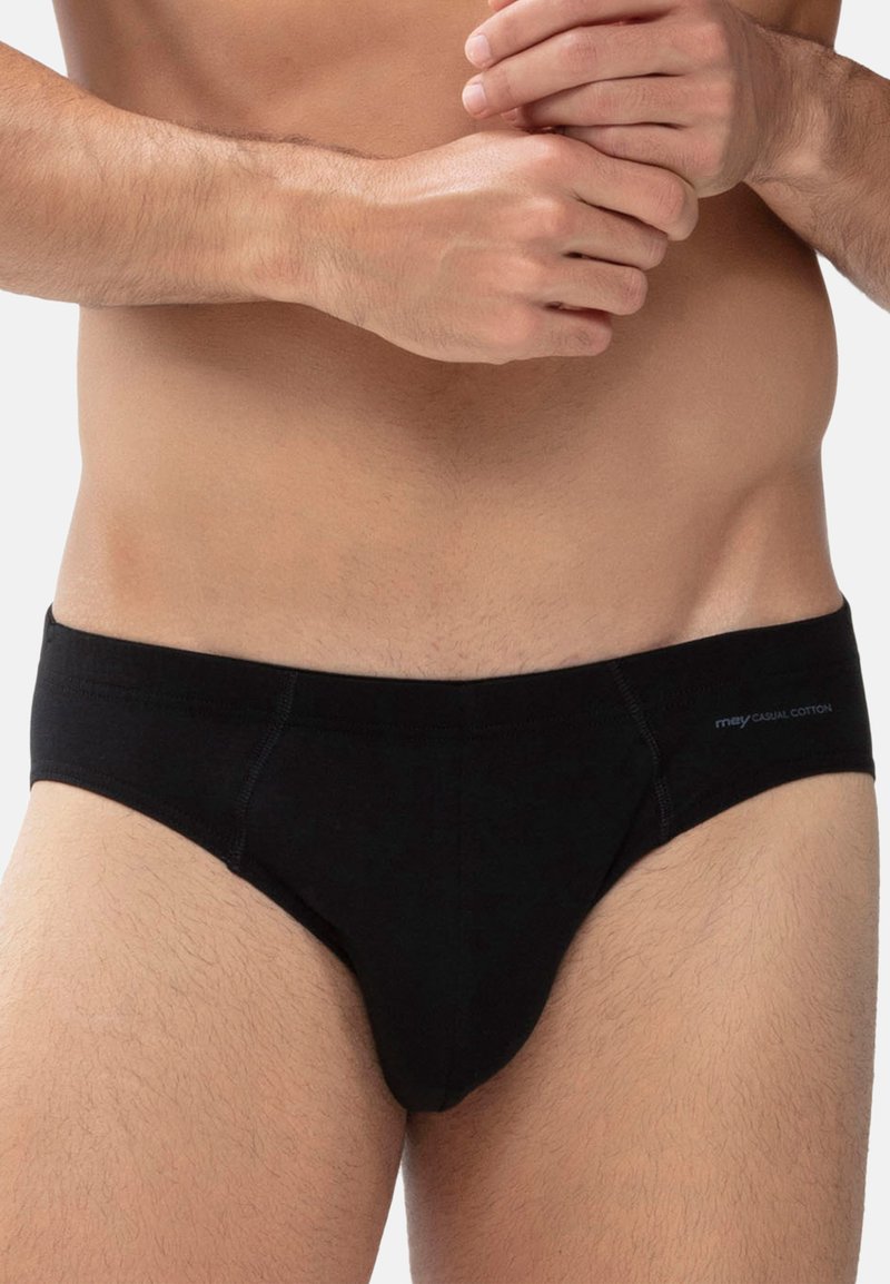 Buste masculin portant un slip en coton noir avec l’étiquette « mey casual cotton », mains croisées à la taille, fond neutre.