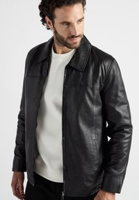 Zwarte leren jack met een rits aan de voorkant, kraag en zijzakken. Gladde textuur en slim fit, gedragen over een witte trui.