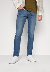 Osoba v světle modrých slim-fit džínách, bílých teniskách, olivově zelené košili a béžové bundě, stojící s jednou rukou v kapse.