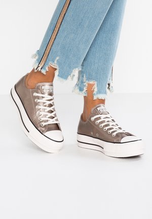 Person iført metallisk bronze Converse All Star plateau sneakers og slidte lyseblå jeans med sidelommer, stående på hvidt gulv.