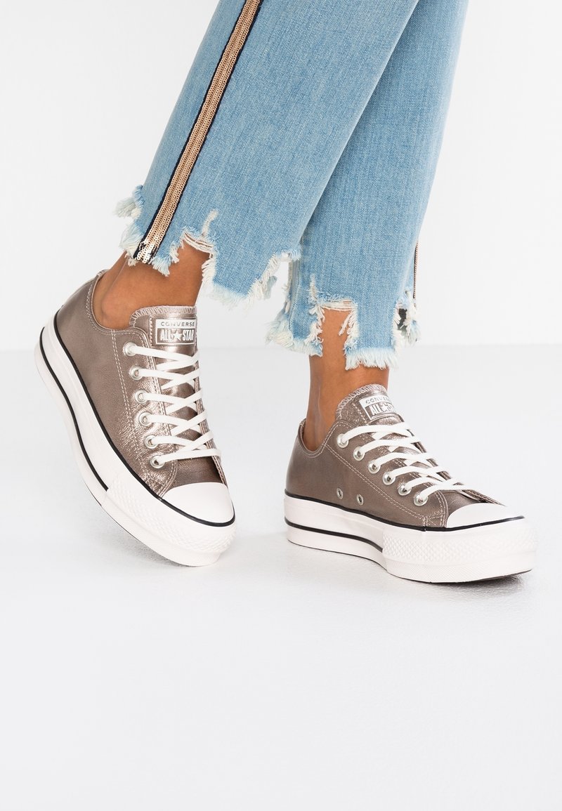 Personne portant des baskets à plateforme Converse All Star en bronze métallique et un jean bleu clair effiloché avec des bandes latérales, debout sur un sol blanc.