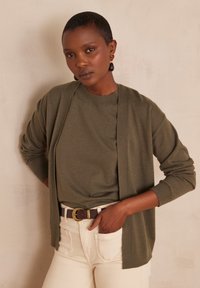 Cardigan leggero verde oliva sopra una t-shirt abbinata, abbinato a jeans color crema e cintura marrone. Orecchini pendenti di rilievo.