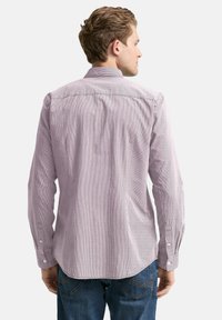 Ein Mann, der mit dem Rücken zur Kamera steht, trägt ein helllila gestreiftes Langarmhemd und blaue Jeans vor einem schlichten, hellen Hintergrund.