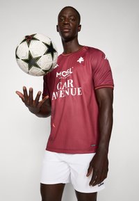 Rød sportsdrakt med hvite detaljer og logoer, båret av en person som holder en svart og hvit fotball, sammen med hvite shorts.