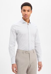 Camicia a maniche lunghe con base bianca caratterizzata da un motivo geometrico multicolore, abbinata a pantaloni grigio chiaro. Colletto standard, davanti con bottoni.