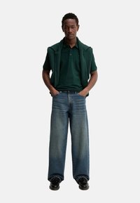 Camisa polo verde, jeans azules de corte holgado y zapatos negros. Presenta un cuello con solapa y un bolsillo frontal, con un ajuste relajado y denim descolorido.