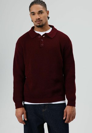 Man met ingevlochten haar, gekleed in een bordeauxkleurige gebreide trui over een wit overhemd en donkere spijkerbroek, staand tegen een effen grijze achtergrond.