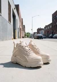 Kraftige beige sneakers med tykke såler placeret på en byfortov med bygninger og parkerede biler i baggrunden under en klar himmel.