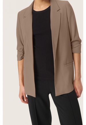 Kobieta ubrana w beżową marynarkę z marszczonymi rękawami, czarny prążkowany top i czarne spodnie na jednolitym tle.