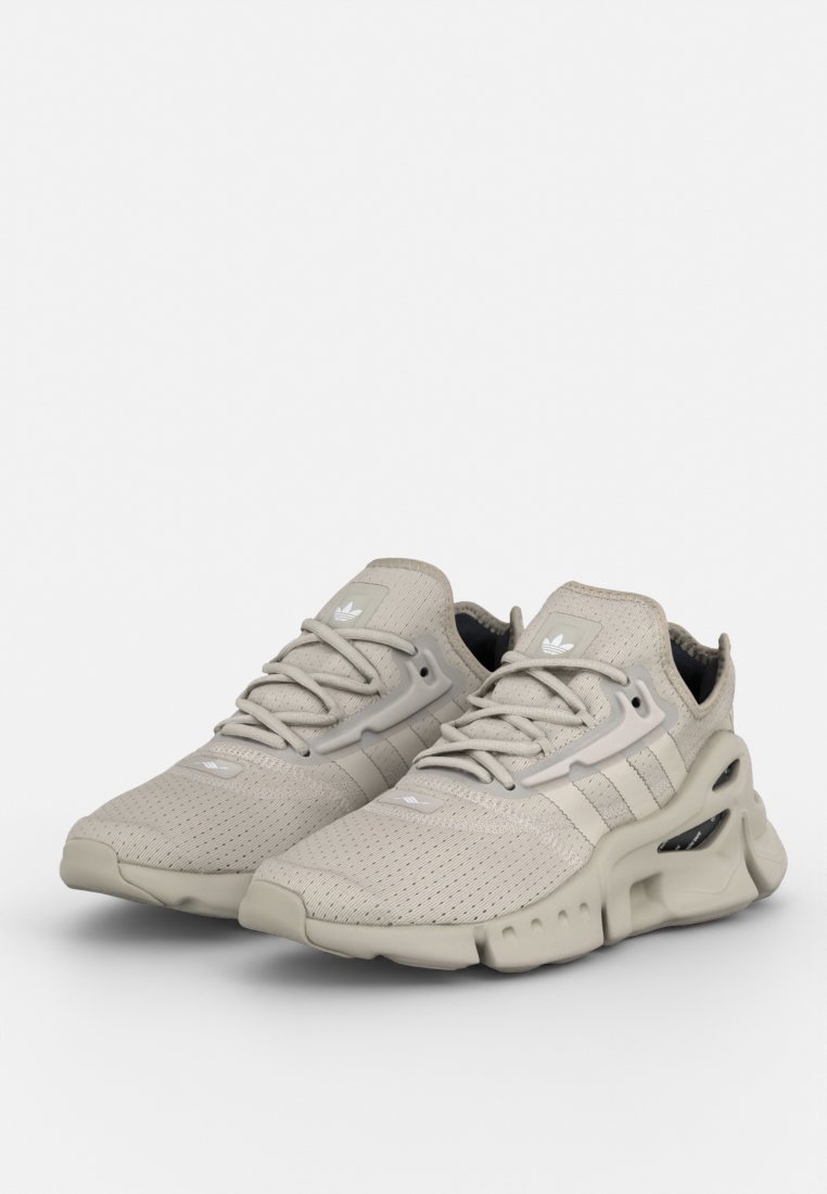 Zapatillas deportivas beige con parte superior de malla, detalles texturizados y una suela moldeada con recortes. Marca de tres rayas en gris claro.