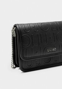 Clutch de cuero negro texturizado con logo en relieve, correa de cadena plateada y cierre de solapa. Forma rectangular con líneas y detalles limpios.