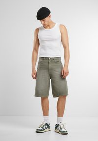 Shorts in denim verde con una vestibilità ampia, abbinati a una canotta bianca e a un berretto lavorato a maglia nero. Calze bianche e sneakers verde e bianche completano il look.