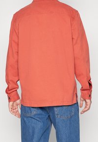 Orange bomulljacka med rak skärning, långa ärmar, knäppta manschetter och sidofickor, tillsammans med blå denimjeans.