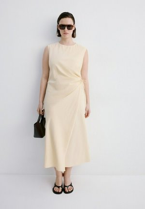 Mujer con vestido midi sin mangas color crema, sandalias negras, gafas de sol oscuras, sosteniendo un pequeño bolso negro, de pie contra una pared blanca.