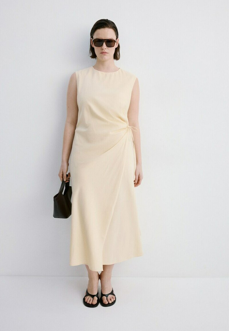 Femeie purtând o rochie midi crem fără mâneci, sandale negre, ochelari de soare închiși la culoare, ținând o geantă mică neagră, stând în fața unui perete alb.