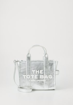 Hopeinen metallinhohtoinen kassi, jossa on kaksinkertaiset yläkahvat, irrotettava olkahihna ja rohkea valkoinen typografia, jossa lukee "THE TOTE BAG MARC JACOBS."