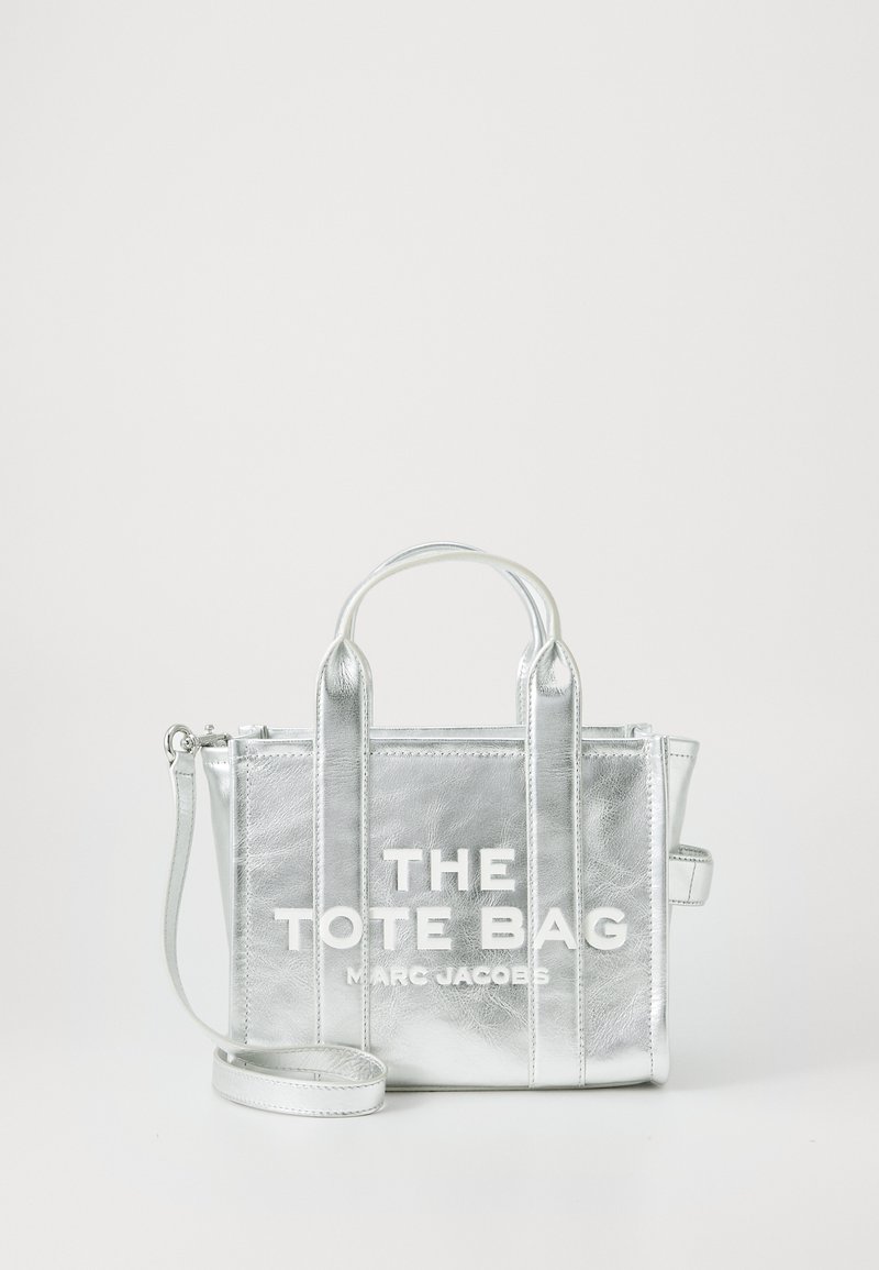 Sudraba metāliskā somiņa ar divām augšējām rokturiem, noņemamu plecu siksnu un izteiksmīgu baltu tipogrāfiju, kas lasāma "THE TOTE BAG MARC JACOBS."