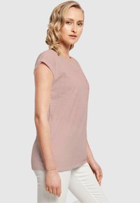 Lys rosa T-shirt med korte ærmer, rund halsudskæring og en diskret frontlomme, som bæres med hvide bukser. Glat, letvægtsstof.