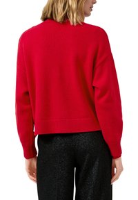 Roter, gestrickter Pullover mit cropped Design, überschnittenen Schultern und gerippten Bündchen. Getragen mit glänzenden schwarzen Hosen, die einen strukturierten Stoffkontrast zeigen.