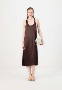 Brun uten ermer maxi kjole med rund hals og knelengde. Modellen holder en liten hvit veske med teksturert overflate.