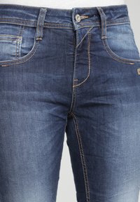 Detailní záběr střední části těla ve modrých džínech s vybledlými místy a oranžovým prošíváním, zapnutými v pase.