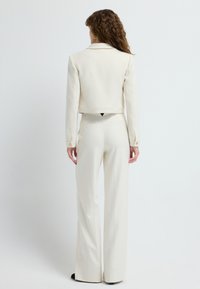 Elfenben farvet cropped jakke med klippet krave og lange ærmer, parret med højtaljede, flagrende bukser i matchende stof. Glat tekstur, minimale accenter.