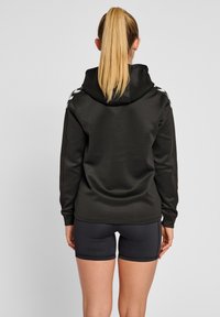 Schwarzer Kapuzenpullover aus glattem Material, mit weißen Akzenten an den Schultern, kombiniert mit schwarzen Shorts. Einfaches Design, lässige Passform.