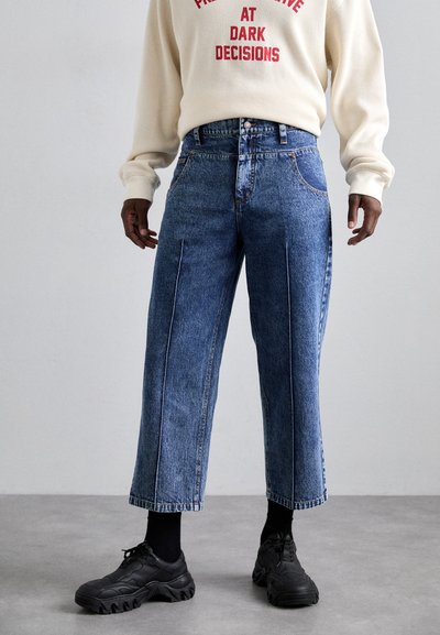 Jeans in denim azzurro chiaro a gamba larga con cuciture bianche e spacchi laterali. Abbinati a sneaker nere e a un maglione color crema con testo rosso.