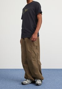 Černé tričko Nike s logem, béžové cargo kalhoty s bočními kapsami a šedé sportovní boty s černými akcenty, stojící na modrém koberci.