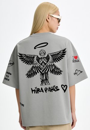 Persoon met lang blond haar die een zonnebril draagt en een grijs T-shirt met een zwarte engel grafiek en de tekst "MIRA PARIS" op de rug.