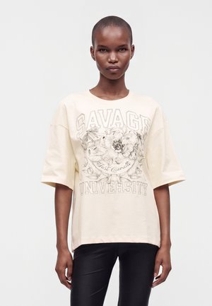 Frau mit kurzen Haaren trägt ein übergroßes beiges T-Shirt mit floralem und textlichem Design mit der Aufschrift "Savage University Just Cavalli."