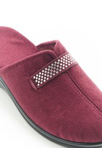 Pantofole slip-on bordeaux realizzate in un morbido tessuto con una texture simile alla pelle scamosciata, caratterizzate da una banda decorativa di strass sulla parte anteriore.