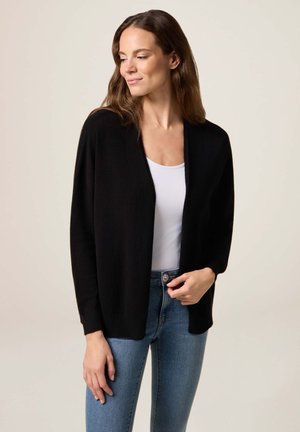Schwarze offene Strickjacke aus weichem Material, mit langen Ärmeln und einem eleganten, lässigen Schnitt. Kombiniert mit einem weißen Oberteil und blauen Jeans.