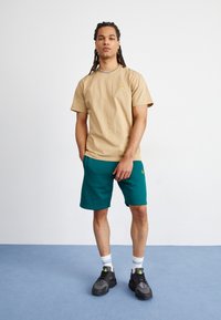 Beige Baumwoll-T-Shirt mit einem kleinen gelben Logo auf der Brust, kombiniert mit türkisfarbenen Shorts. Das Modell trägt schwarze Turnschuhe und weiße Socken mit blauen Streifen.