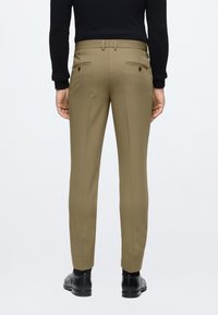 Khaki op maat gemaakte broek met een slim fit, voorzien van achterzakken en riemlussen, gecombineerd met zwarte schoenen. Zachte stoftextuur.