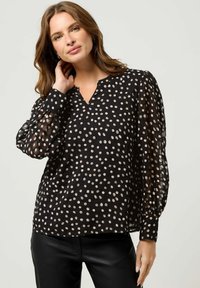 Blusa nera a pois con scollatura a V e maniche a sbuffo in tessuto trasparente. Presenta pois color crema disposti in un motivo sparso su tutto il tessuto.