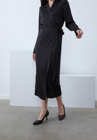 Robe portefeuille noire en tissu lisse, dotée de manches longues, d'une ceinture à la taille et d'une longueur mi-mollet. Associée à des chaussures à talons hauts noires.