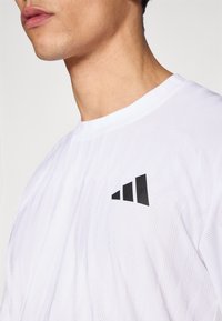 Camiseta blanca de manga corta con patrón texturizado; presenta un detalle de logo negro en el lado izquierdo. Tejido ligero y transpirable.