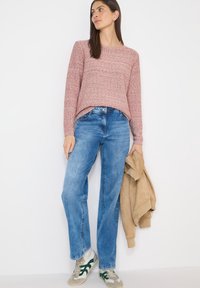 Femme en pull rouge à motifs et jean bleu tenant une veste beige, portant des baskets blanches avec des rayures vertes, debout devant un fond blanc.