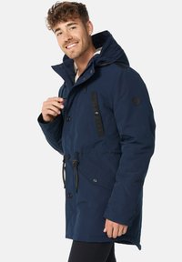Marineblå parka jakke lavet af isoleret stof, med en faux pelsforing i hætten, justerbare snore og lomme med knaplukning.