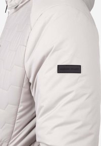 Chaqueta acolchada gris claro con diseño hexagonal texturizado en el hombro y una etiqueta de cuero negro que dice "AMARO JEANS" en el brazo.