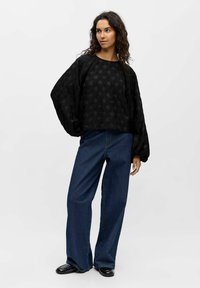 Vrouw met lang krullend haar, gekleed in een losse zwarte textuurblouse, wijdvallende blauwe jeans en zwarte schoenen, staande tegen een effen lichte achtergrond.