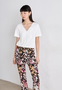 Weißes kurzärmliges T-Shirt mit Schmetterlingsstickerei und tiefem V-Ausschnitt, kombiniert mit bunten gemusterten Hosen in Schwarz, Pink und Orange.