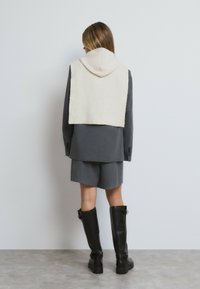 Grauer maßgeschneiderter Blazer über kurzen grauen Shorts, mit einer cremefarbenen Strickkapuze, die über die Schultern drapiert ist, und kniehohen schwarzen Stiefeln. Sanfte Texturen.