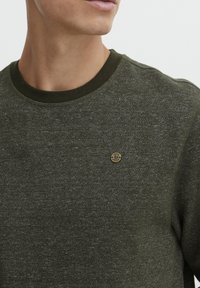 Olijfgroen trui van een gestructureerde stof, ronde halslijn en een klein gouden logootje op de borst. Zacht, casual ontwerp.