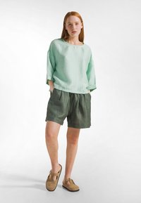 Top in lino verde menta con maniche larghe, abbinato a pantaloncini verde scuro. La modella indossa sandali color cuoio, mostrando una vestibilità rilassata e tessuti morbidi.