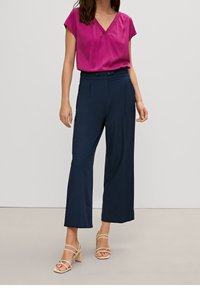 Blouse fuchsia à col en V associée à un pantalon ample bleu marine à jambes larges. Le mannequin porte des talons beige à brides. Tissu lisse, coupe ample et détails plissés.