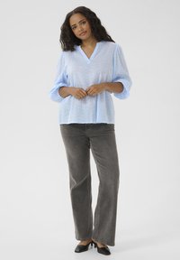 Tunique bleu clair à manches longues avec un col en V, présentant un motif texturé. Associée à un jean large gris et des chaussures à talons noires.