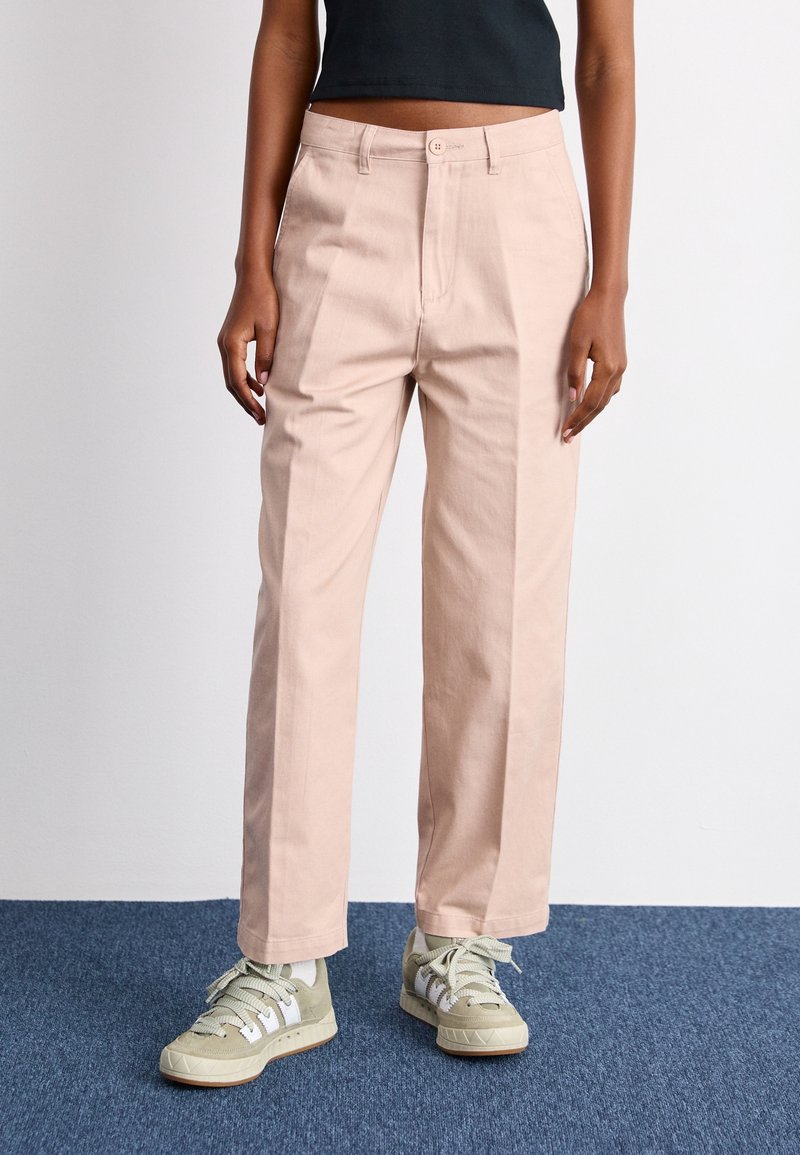 Pantalon taille haute rose clair, coupe droite, avec fermeture avant par bouton. Associé à des baskets vertes.