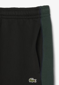 Zwarte joggingsbroek met een groene zijpaneel en blauwe accenten, met een klein geborduurd krokodillenlogo vlakbij de zoom. Gemaakt van een katoenmix.
